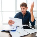 Mann sitzt am Schreibtisch, mit einem Stapel von Zetteln und wirkt ratlos